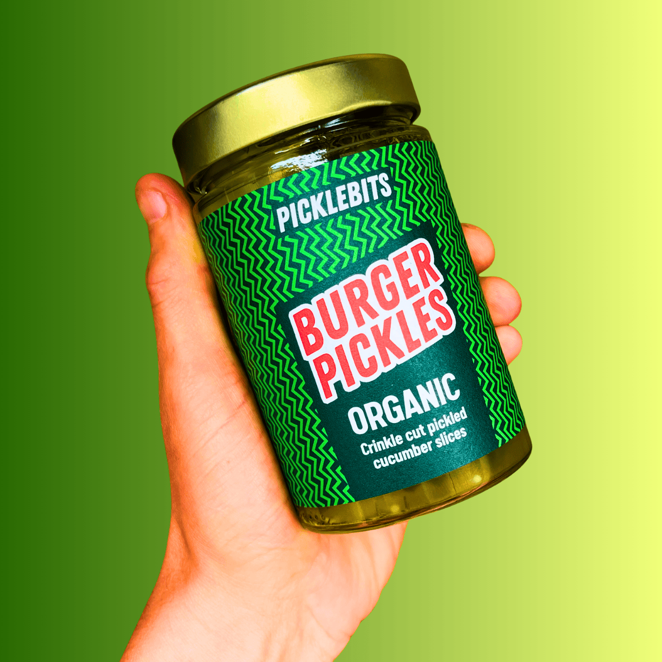 Hand holding a jar of 'Burger Pickles' by PICKLEBITS against a green background